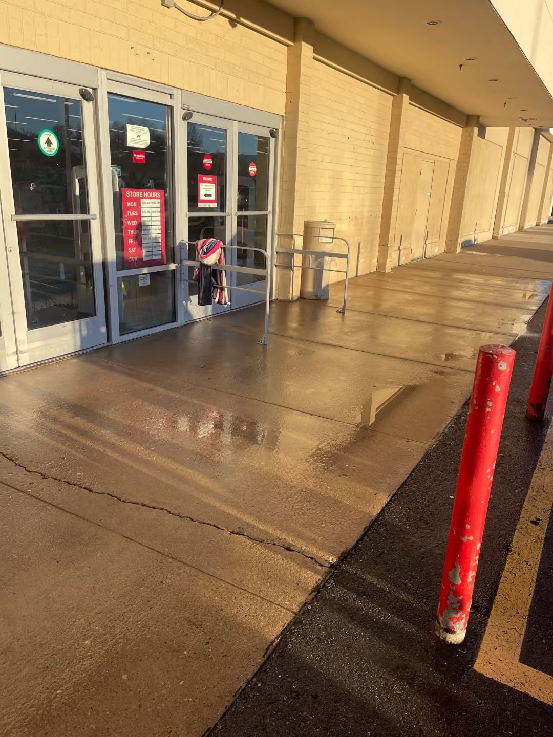Store entrance with wet sidewalk, glass doors, and red bollards in bright sunlight