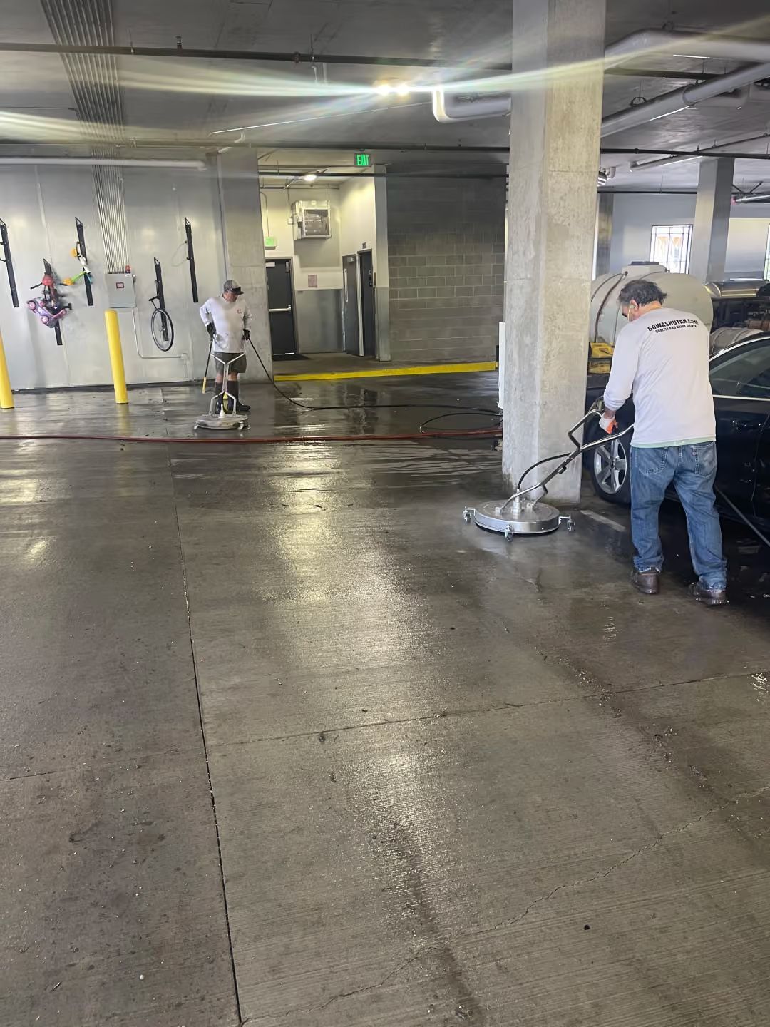 Workers pressure-washing a wet concrete parking garage floor near a yellow curb and hanging gear