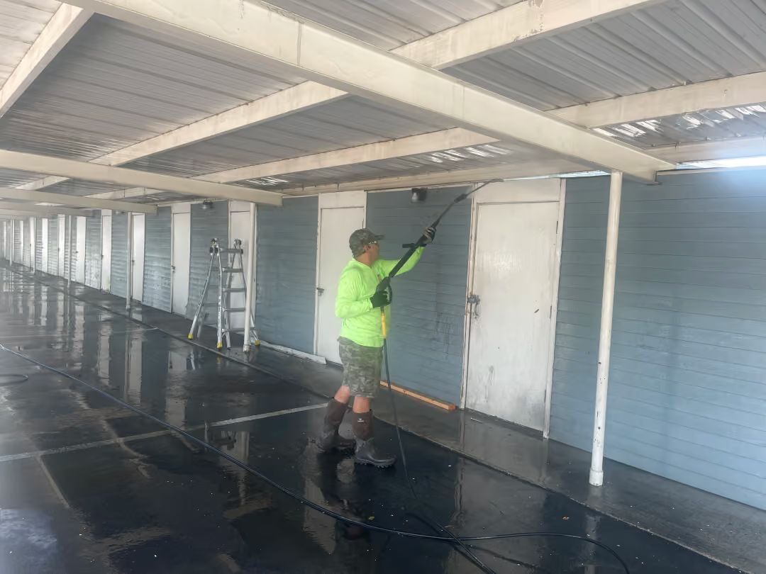 Worker in a neon jacket pressure-washing a corridor with white walls and a wet black floor.
