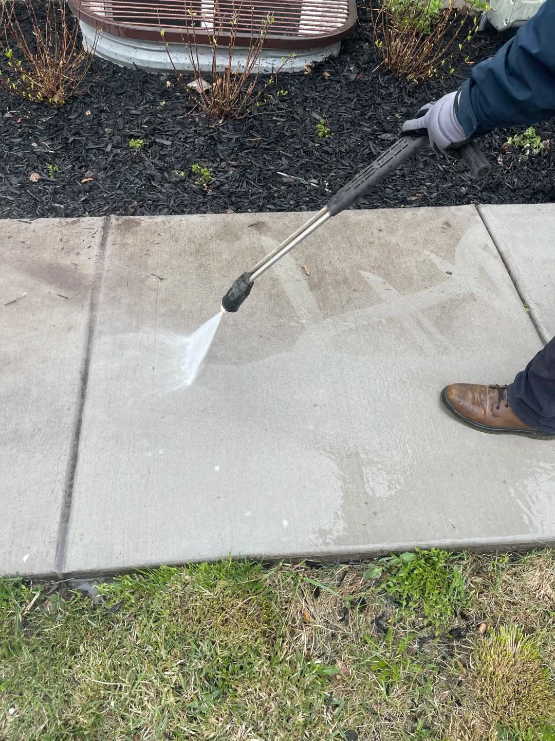 Person pressure-washing a concrete sidewalk beside grass and landscaping.