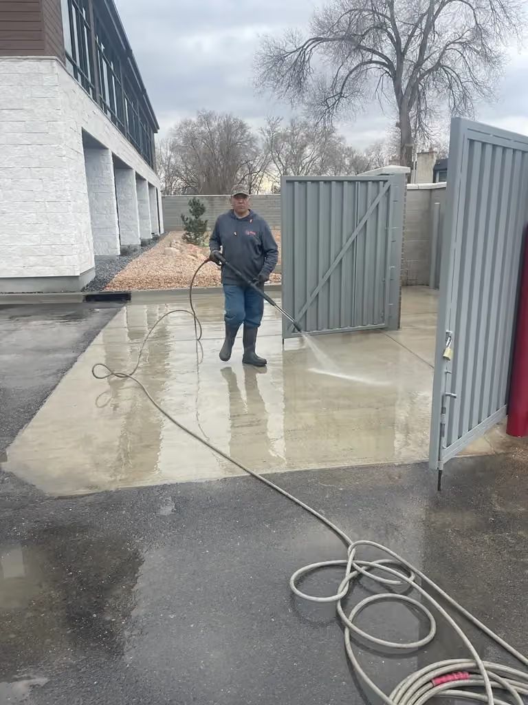 Person washing a concrete walkway with a pressure washer beside a building and open gate.