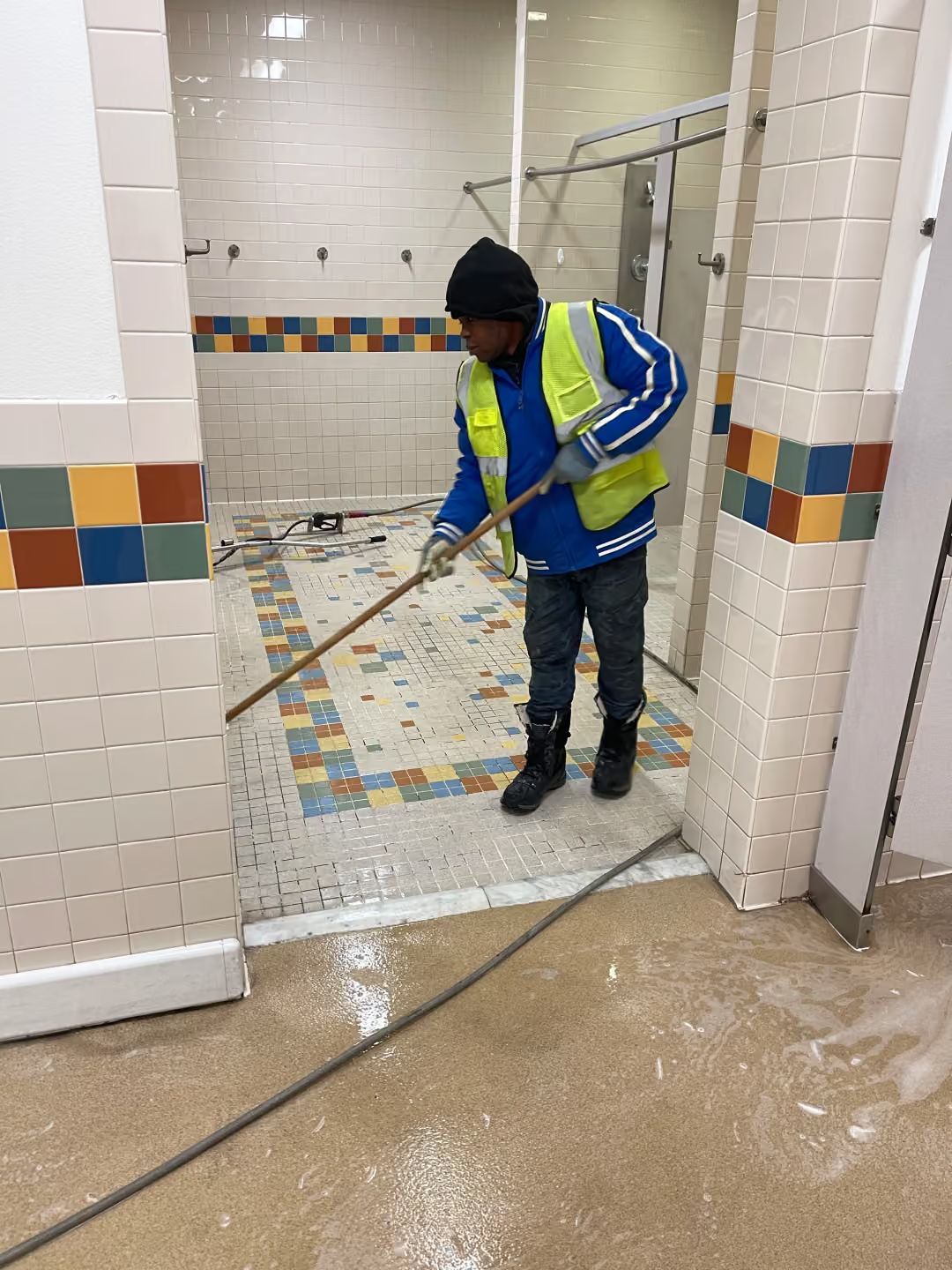 Worker in a blue safety jacket cleaning a tiled restroom floor with a long mop.