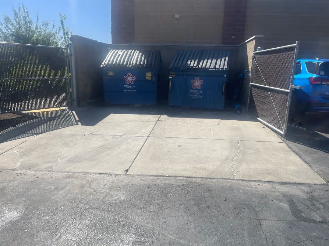 Two blue dumpsters in a fenced concrete alley, with sunlight and shadows