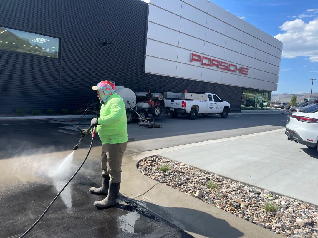 Person pressure washes the sidewalk outside a commercial building with parked trucks.