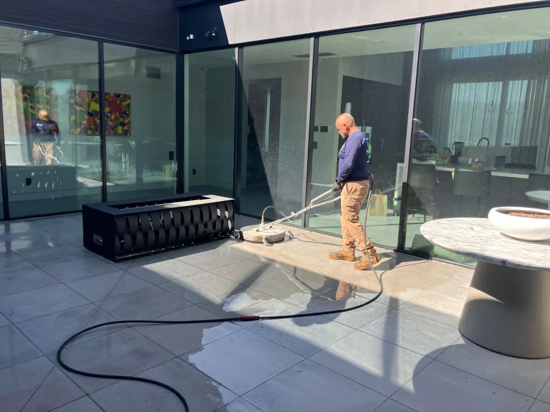 Person pressure-washing a glass patio beside a pool in bright sunlight