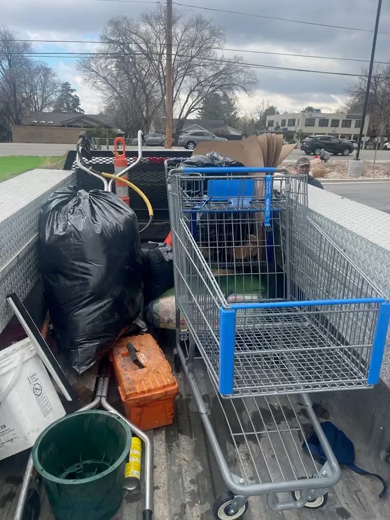 Loaded carts and trash bags on a trailer in a parking lot, with a green bucket and orange crate.