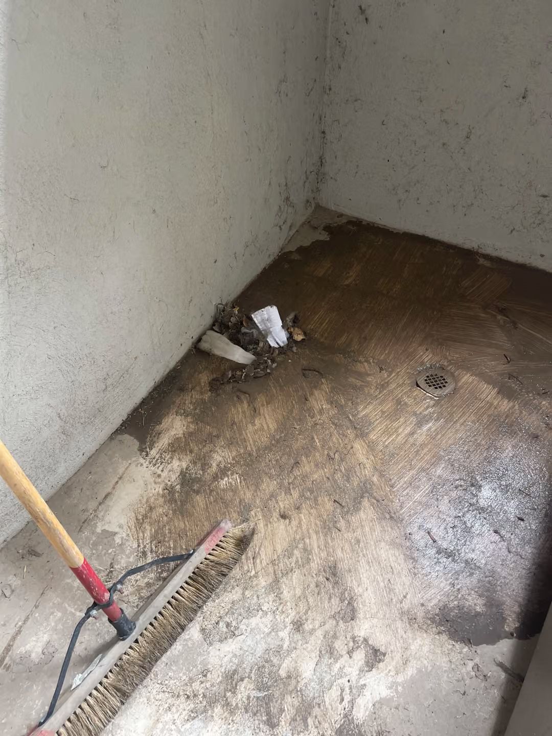 Dirty corner with stained concrete floor, drain, and a broom leaning against the wall