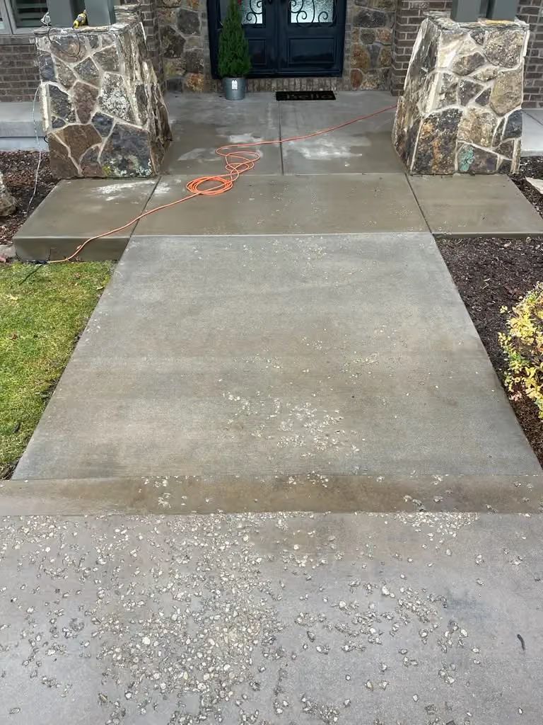 Wet concrete sidewalk leading to a stone-front house entrance with a black door and grassy edges