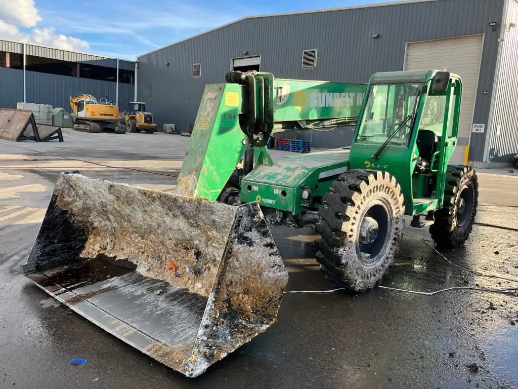 Green telehandler with muddy front bucket parked on wet pavement outside a warehouse