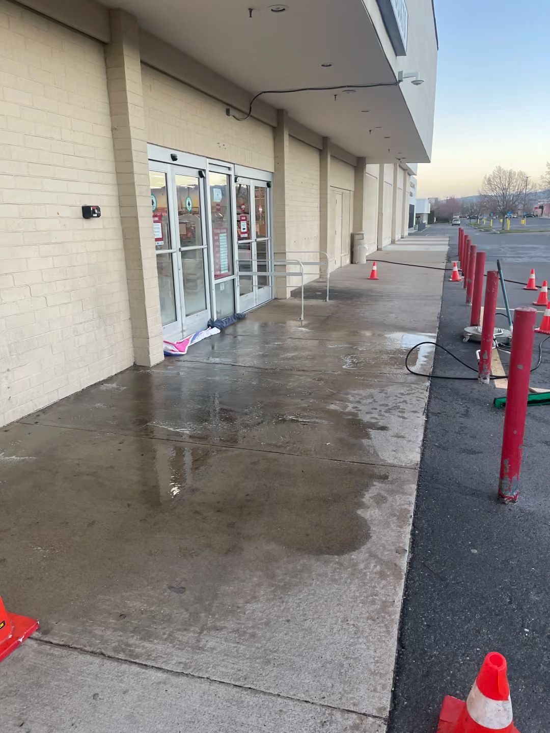Wet storefront entrance with glass doors, red bollards, and orange safety cones along the sidewalk.