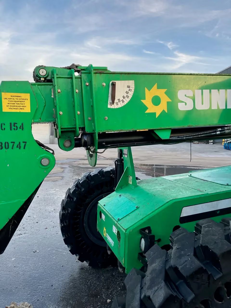 Green Sun agricultural boom lift with large tires parked on wet pavement under a cloudy sky