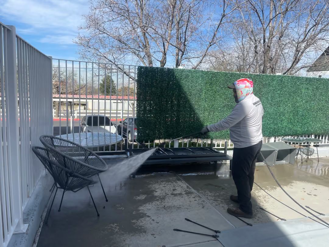 Person in a red hat washes a patio with a hose beside outdoor furniture and a green privacy screen.