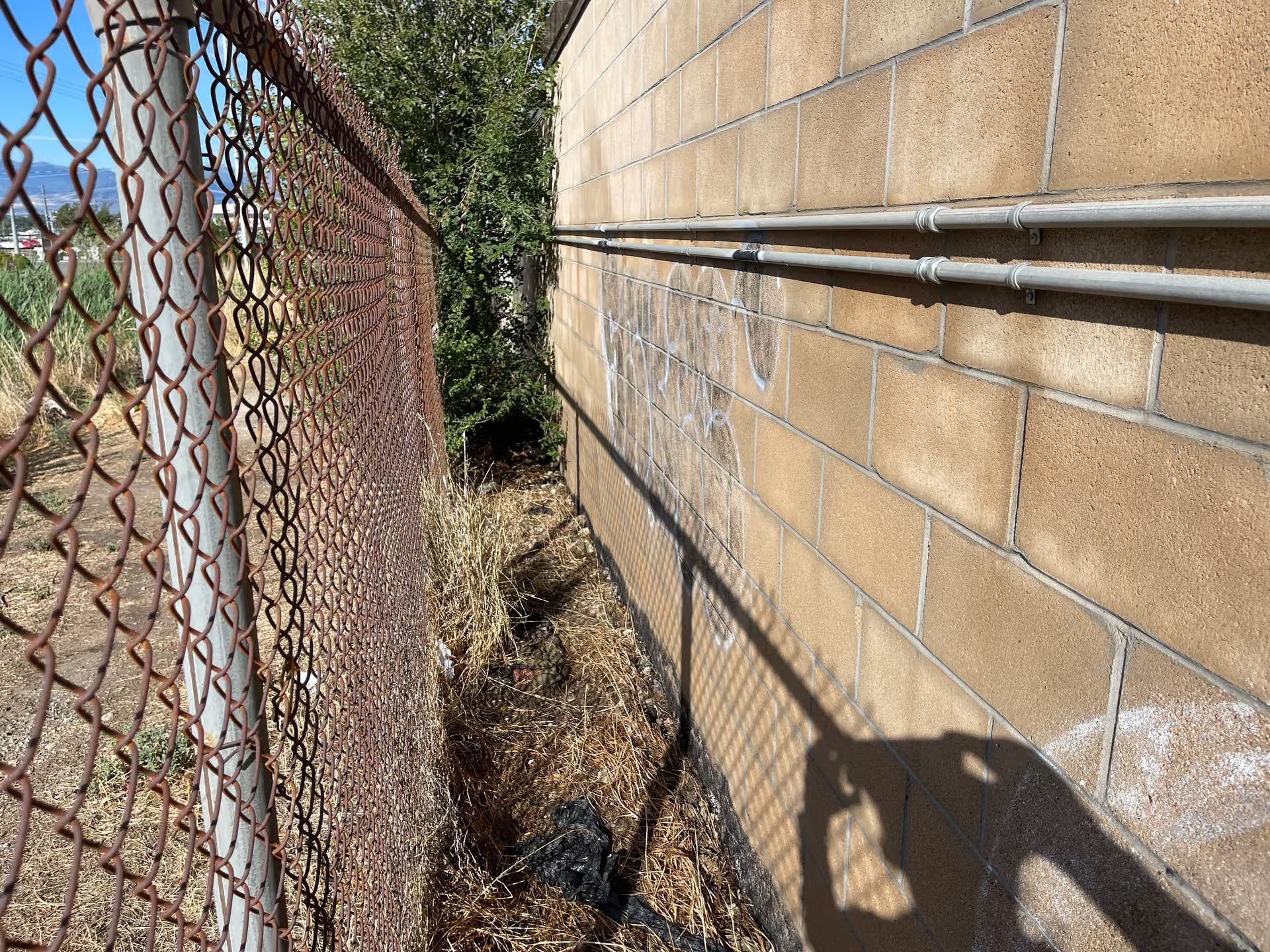 Narrow dirt path between a chain-link fence and brick wall, with pipes running along the wall.