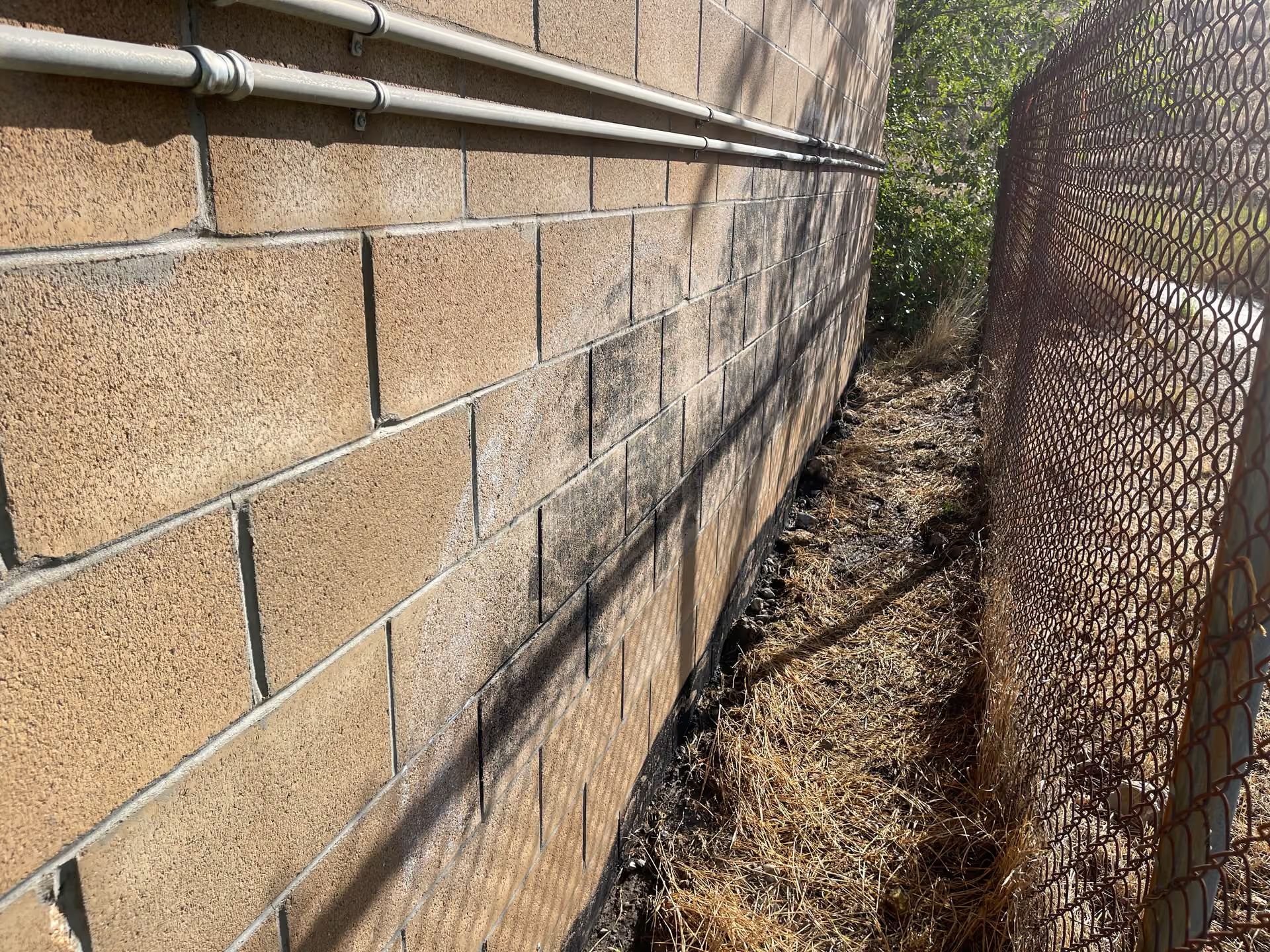 Narrow dirt alley between a tan brick wall and a chain-link fence in bright sunlight