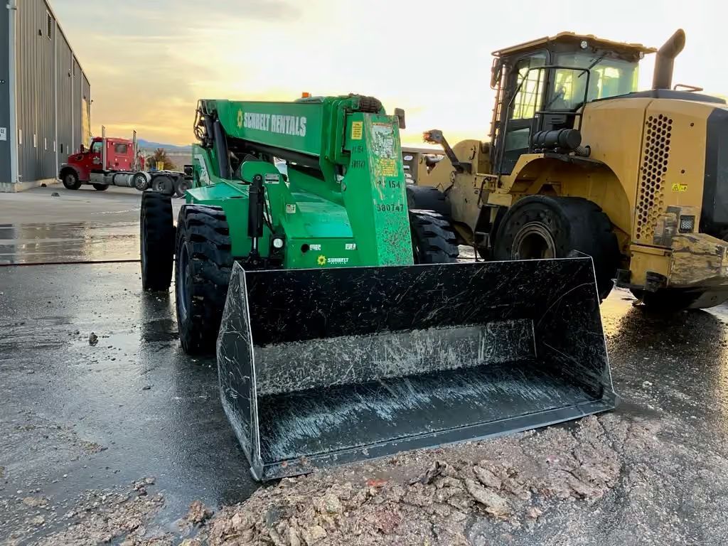 Green telehandler with black bucket beside a yellow loader on a wet construction lot