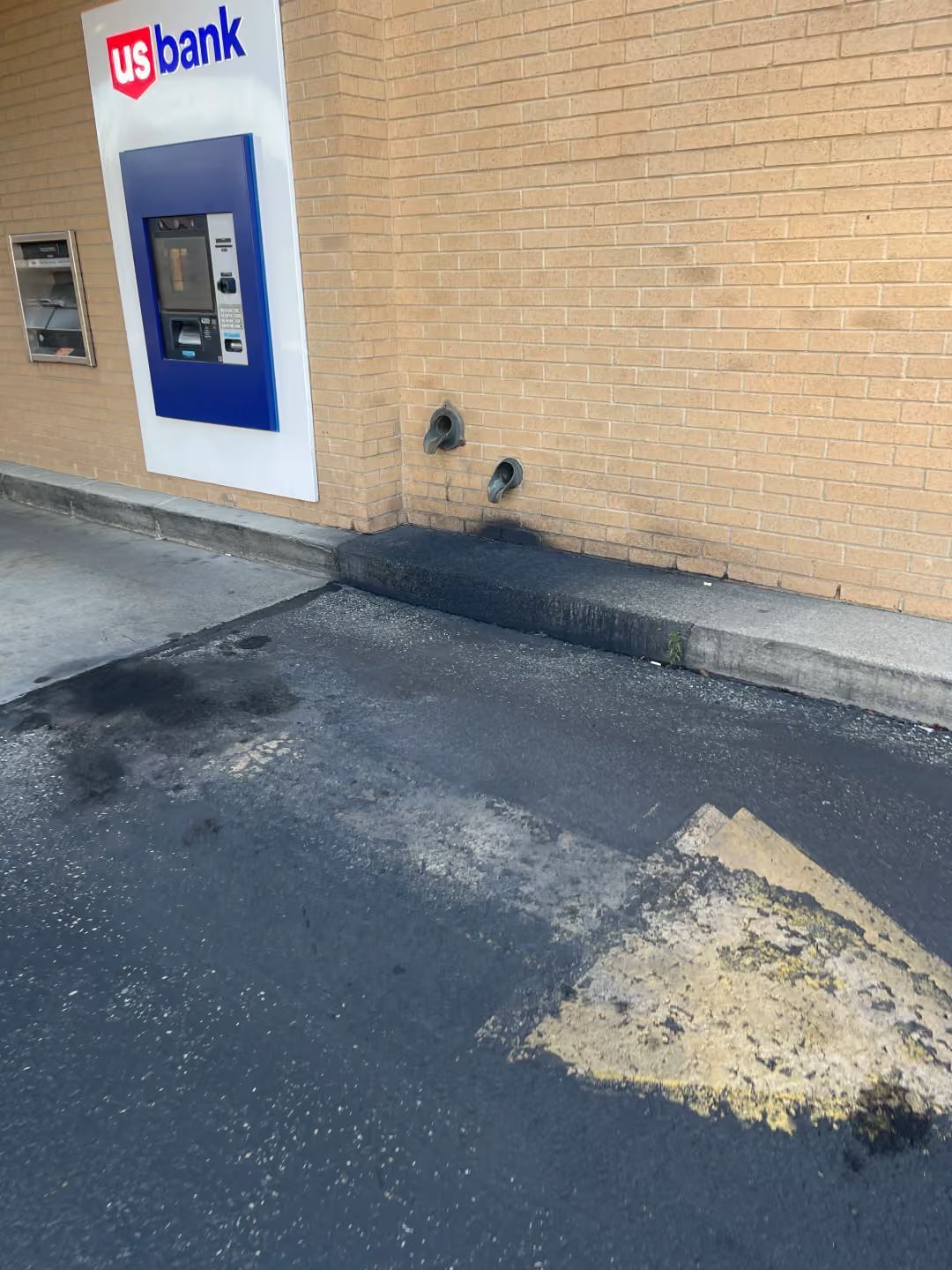 US Bank ATM on a brick wall beside a worn asphalt parking lot with a patch of peeling pavement