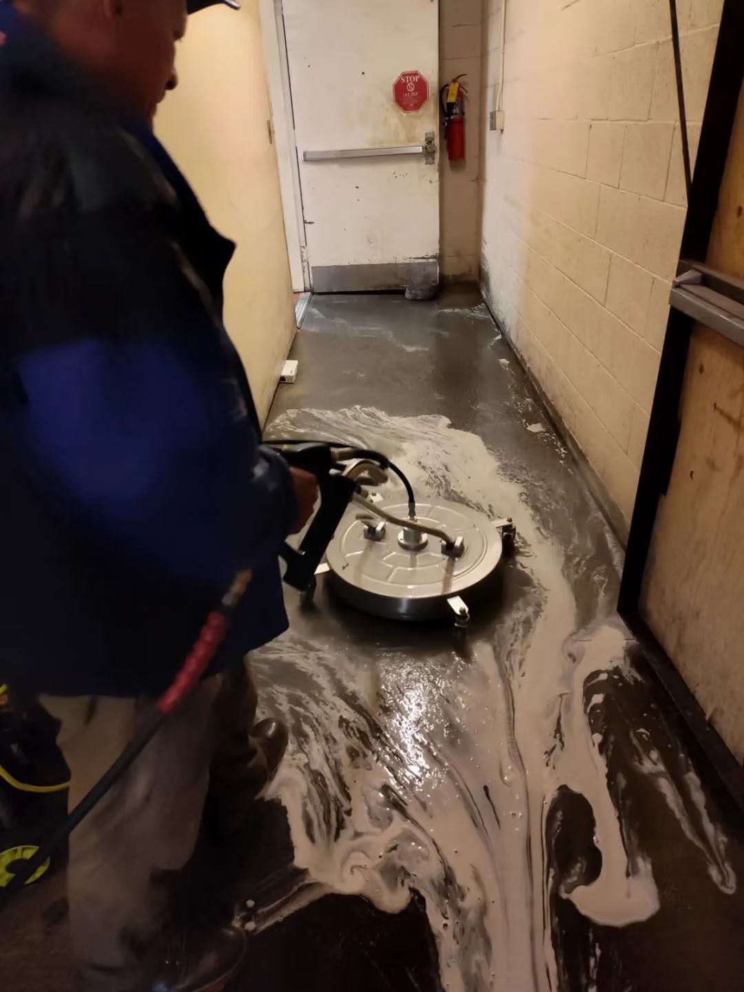 Worker cleaning wet hallway with a floor scrubber, water and foam spreading across the floor.