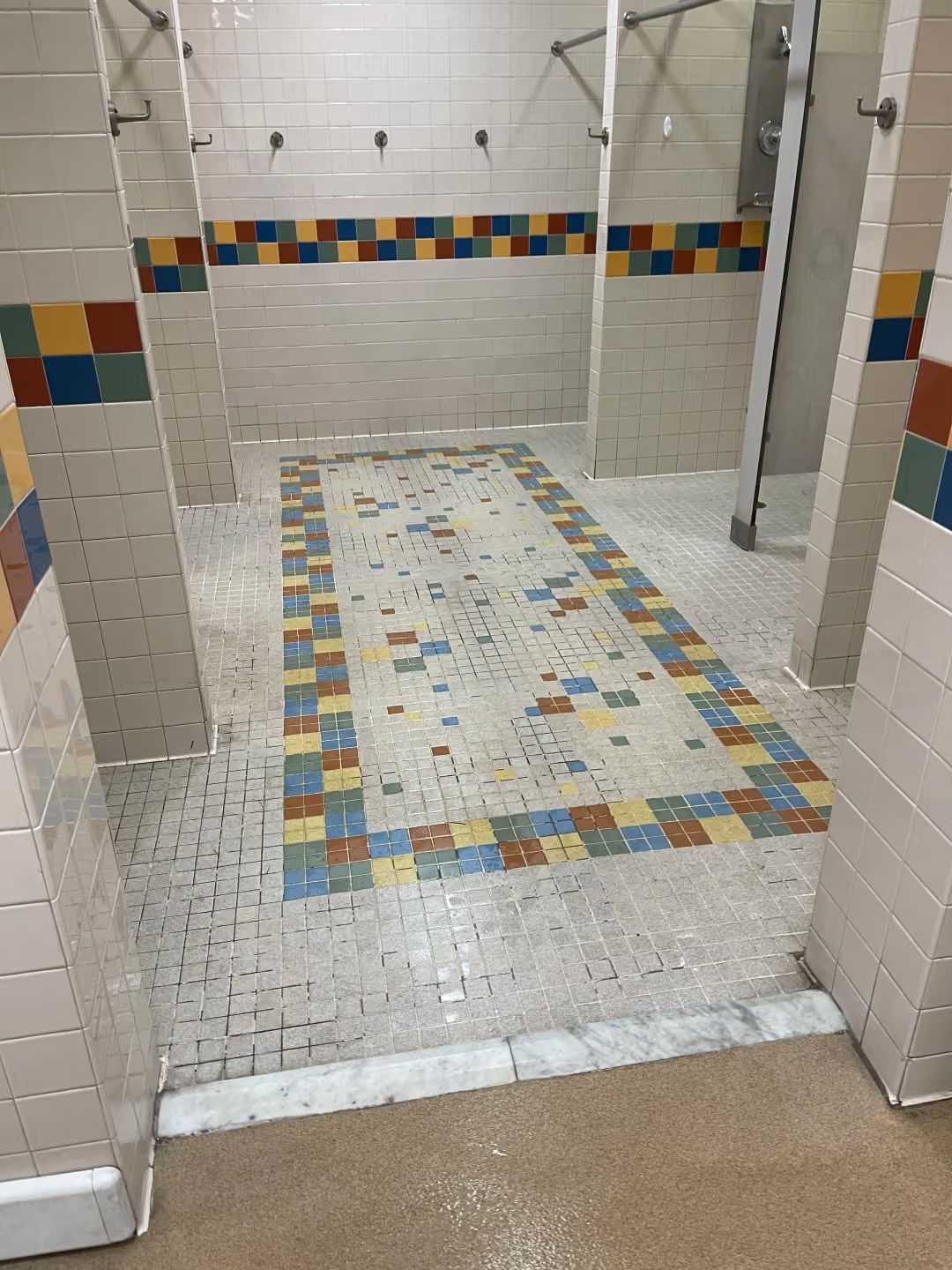 Public shower area with white tiled stalls and a colorful mosaic-tiled floor strip