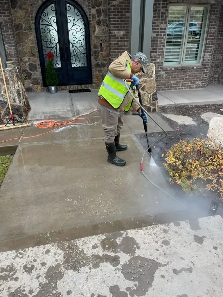 Worker pressure-washing a sidewalk beside a brick house and front door