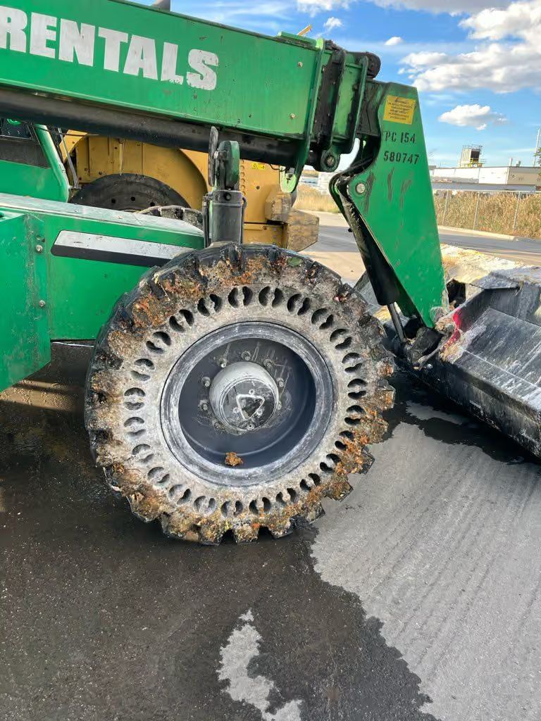 Large green rental loader with a mud-covered wheel on a wet paved road