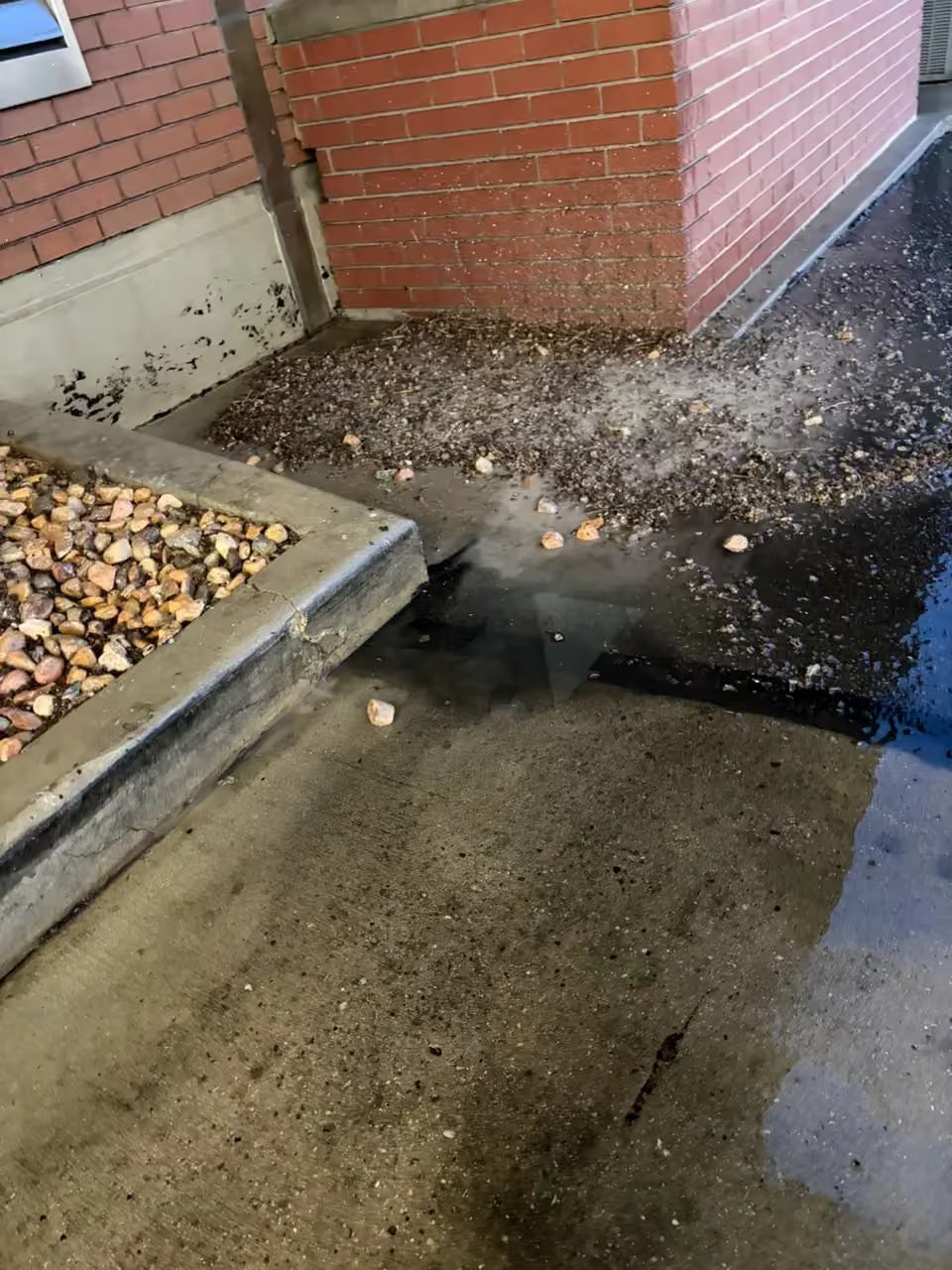 Wet concrete walkway beside a brick building corner, with gravel and a small puddle.
