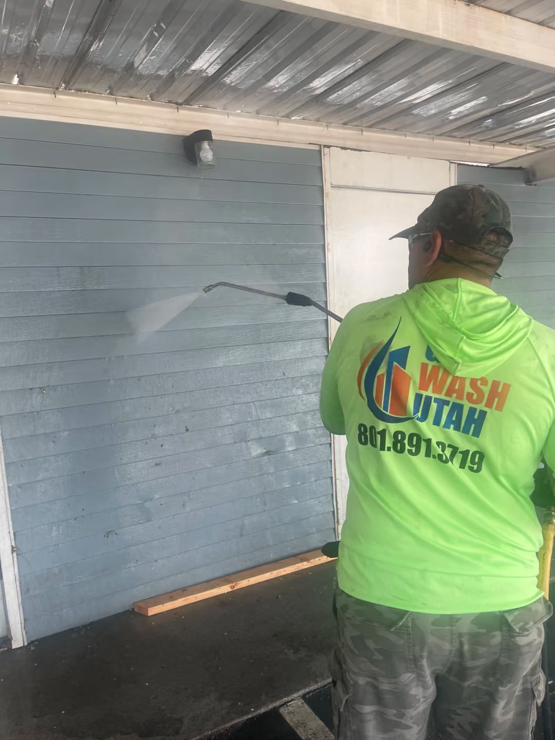 Worker power-washing a blue wall under a metal roof, wearing a bright green shirt.