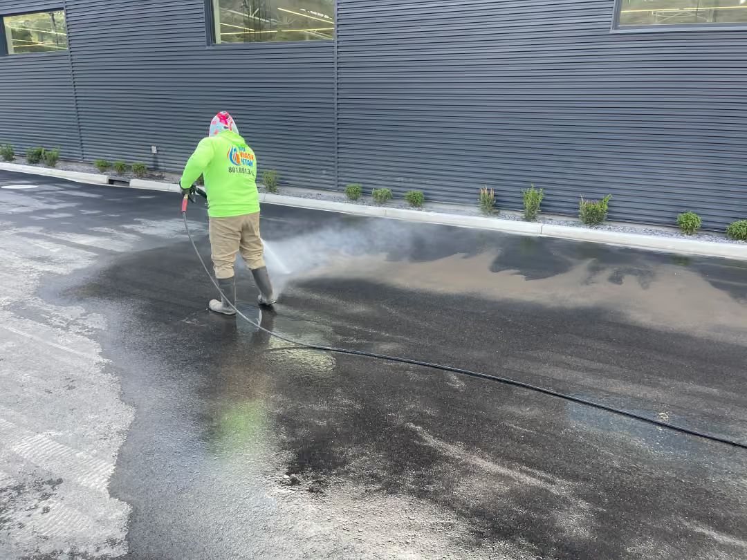 Worker in neon jacket pressure-washing a wet pavement beside a dark corrugated wall.