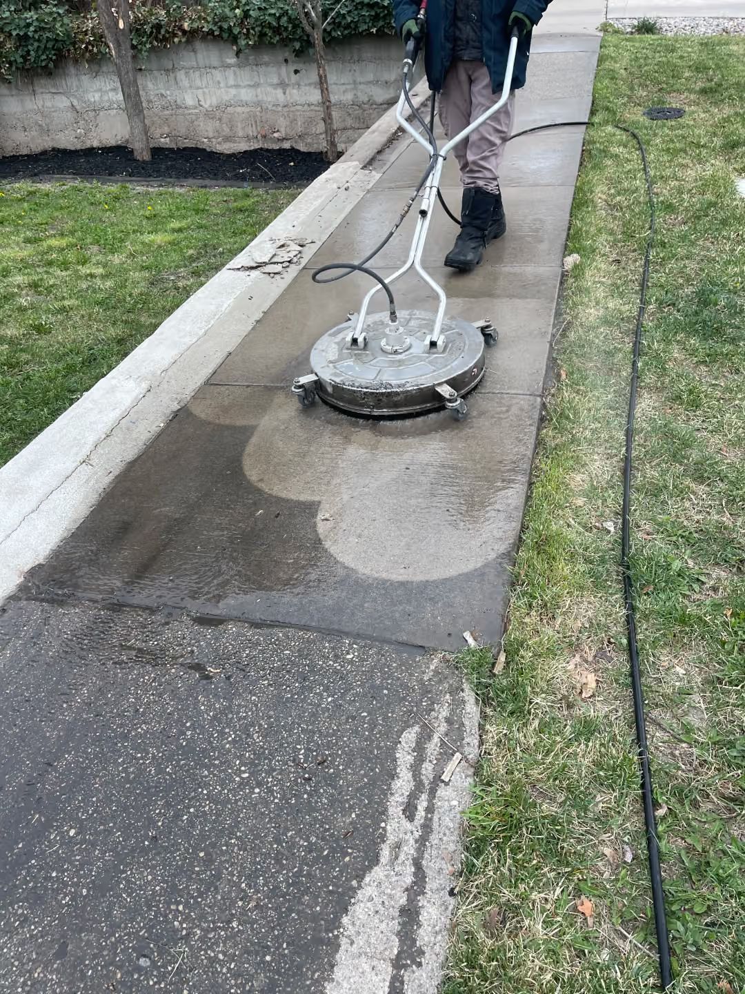 Person pressure-washing a sidewalk beside a grassy strip and curb