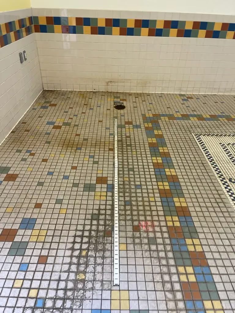 Mosaic-tiled floor in a small tiled room, with dirt stains and a dark drain or hole near the back wall.