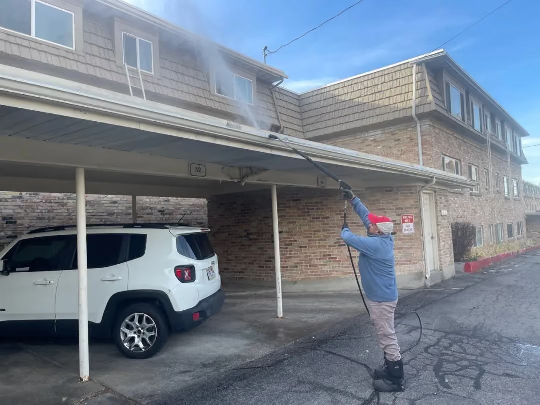 Worker power-washing a carport roof beside a white SUV at an apartment complex