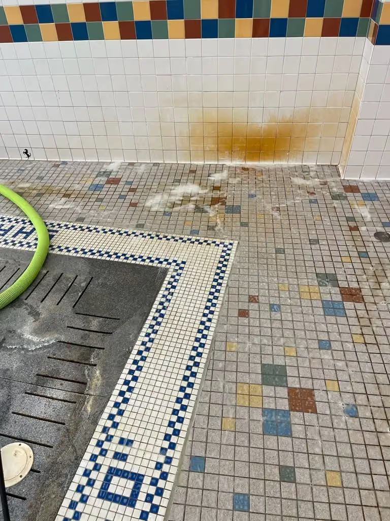 Wet tiled floor beside a shower drain and hose, with an orange stain on the wall.