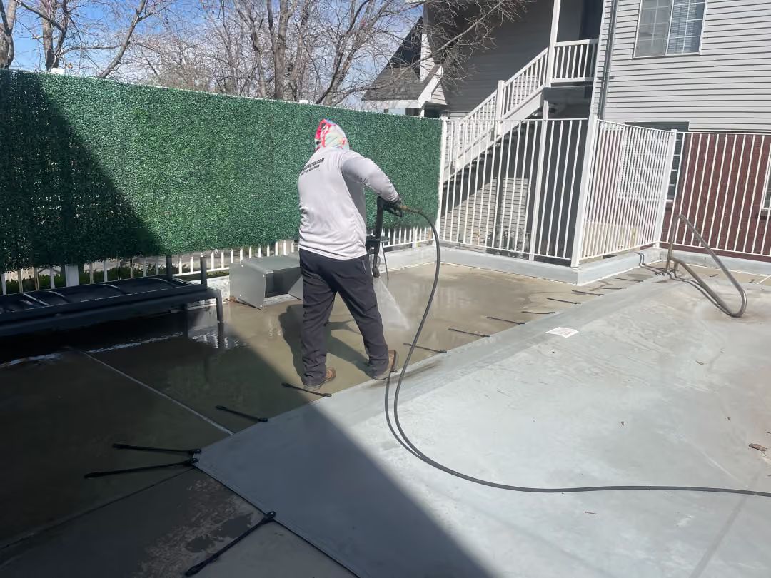 Worker pressure-washing a concrete patio beside a house and stairs on a sunny day