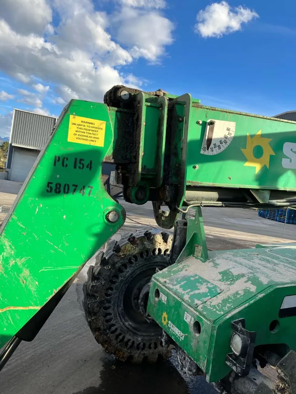 Green construction machine arm and wheel on a paved lot under a blue sky.
