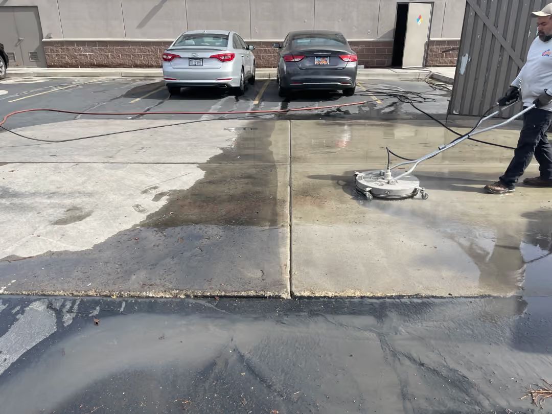Worker pressure-washing a concrete parking lot beside two parked cars and a building