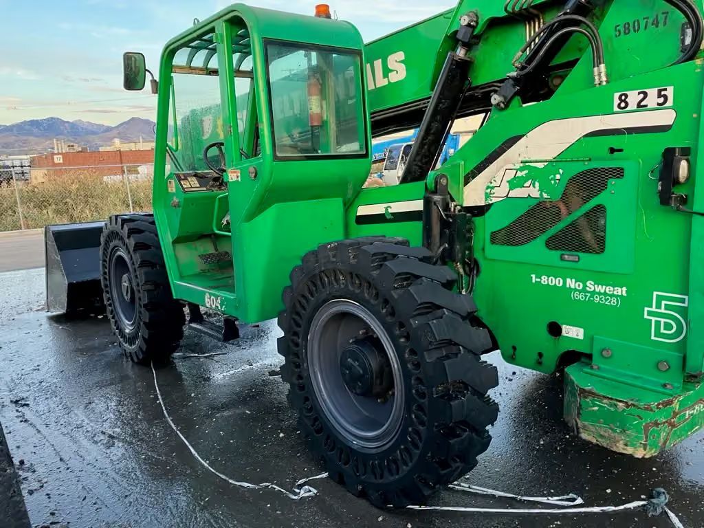 Green logging truck with large front tire on wet pavement, seen from the side outdoors