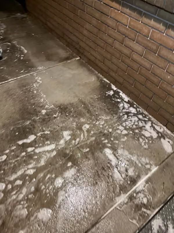 Wet concrete sidewalk beside a brick wall, with white residue and puddles.