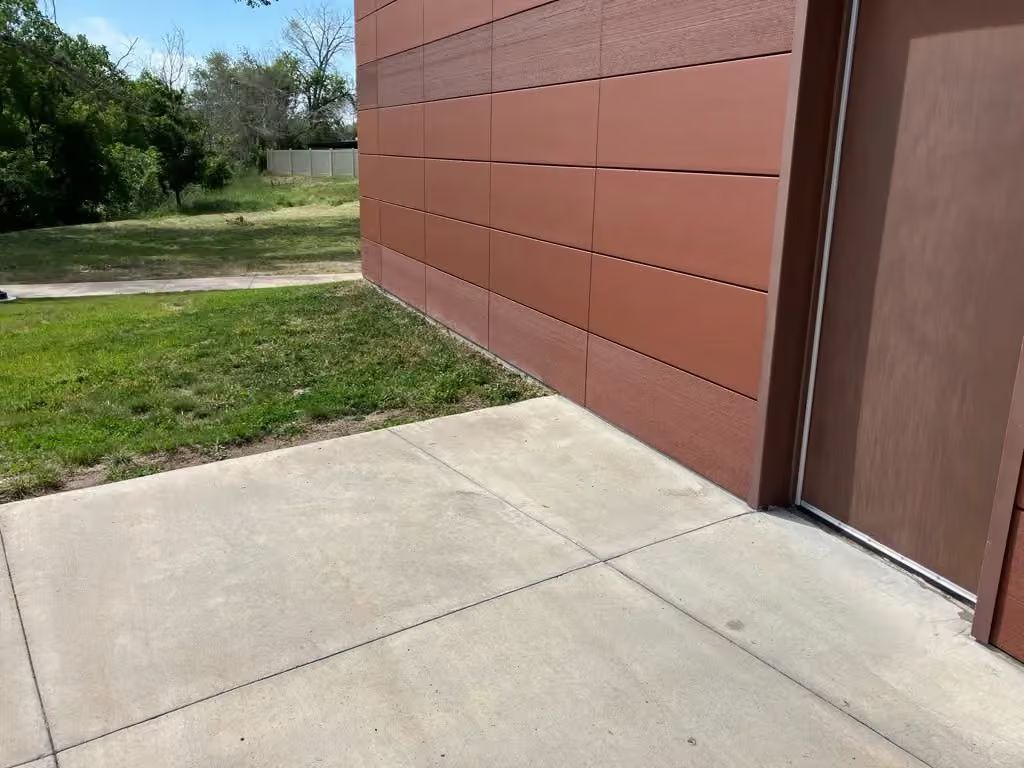 Concrete walkway beside a rust-colored building wall and door, with grass and trees in the background