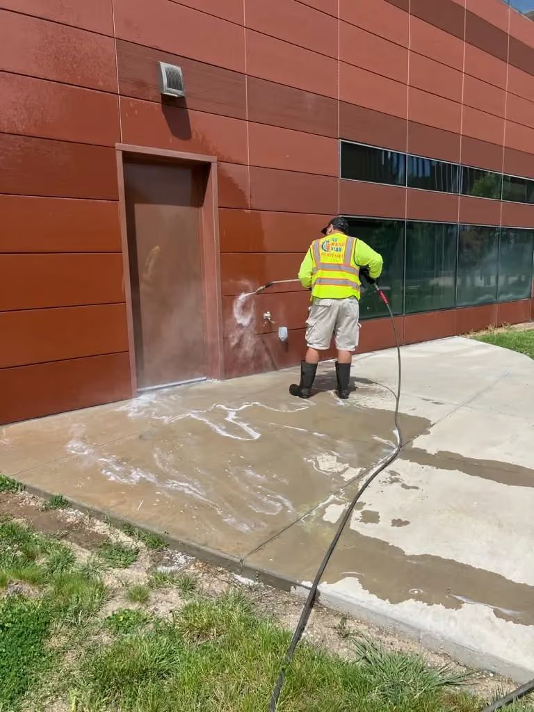 Worker in neon vest pressure-washing a brown building wall and sidewalk.