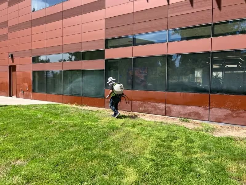 Worker pressure-washing a red building exterior beside a green lawn