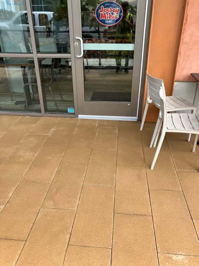 Outdoor storefront entrance with glass doors, tan tile walkway, and stacked white chairs beside a peach wall