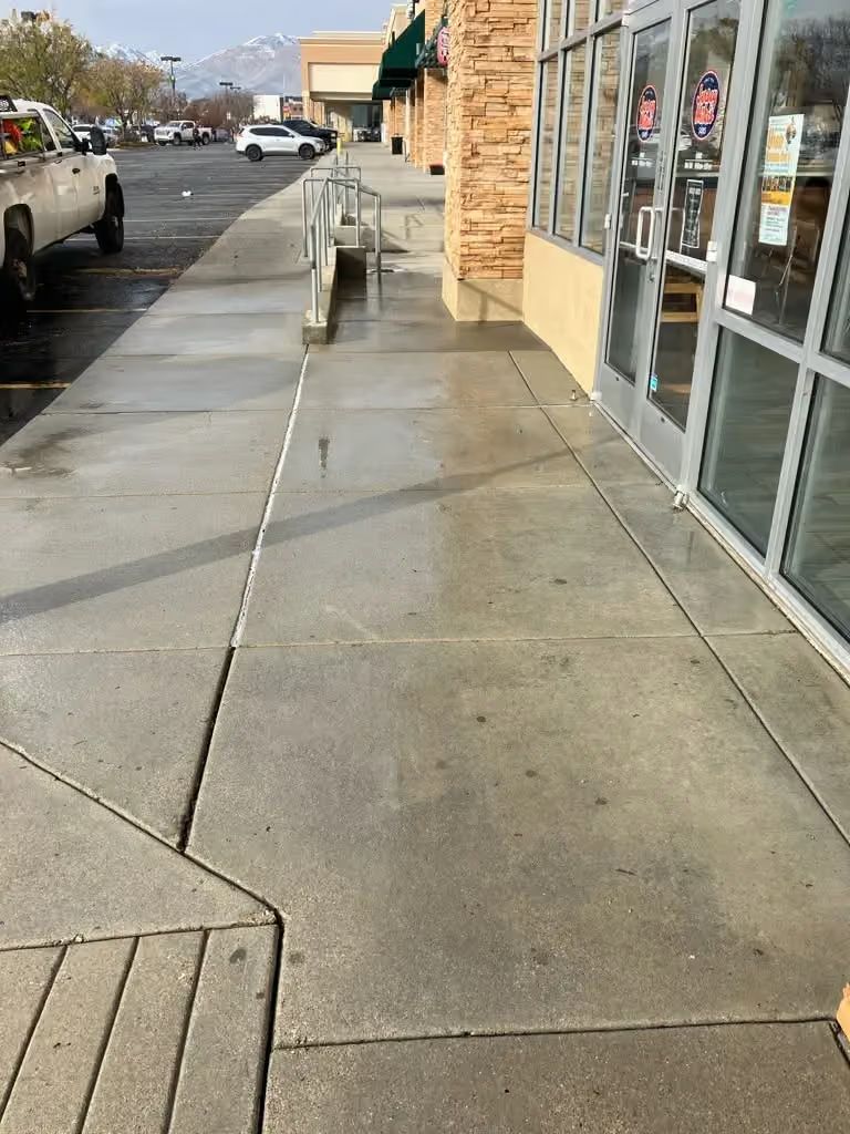 Wet sidewalk beside storefront windows and parked cars on a cloudy day