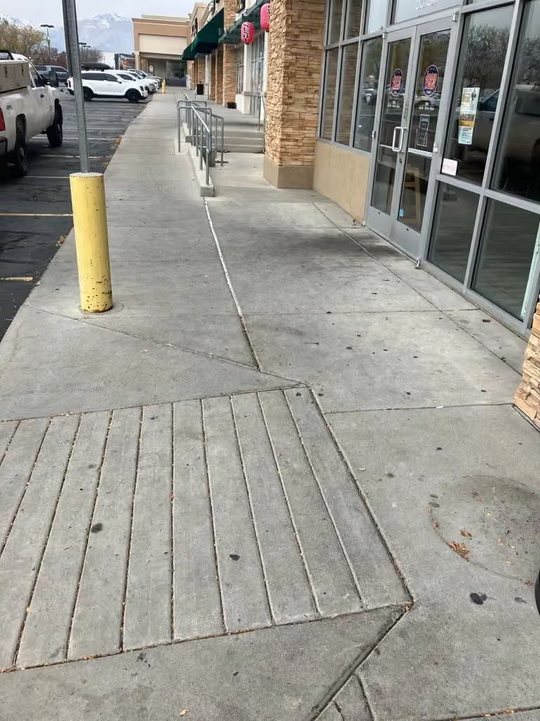 Sidewalk outside a strip mall with storefront windows, a yellow bollard, and an accessible curb ramp