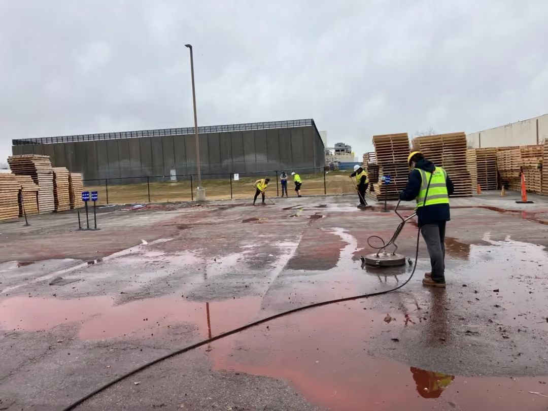 Rainy construction site with workers in yellow vests on a wet red pavement, pallets stacked nearby.