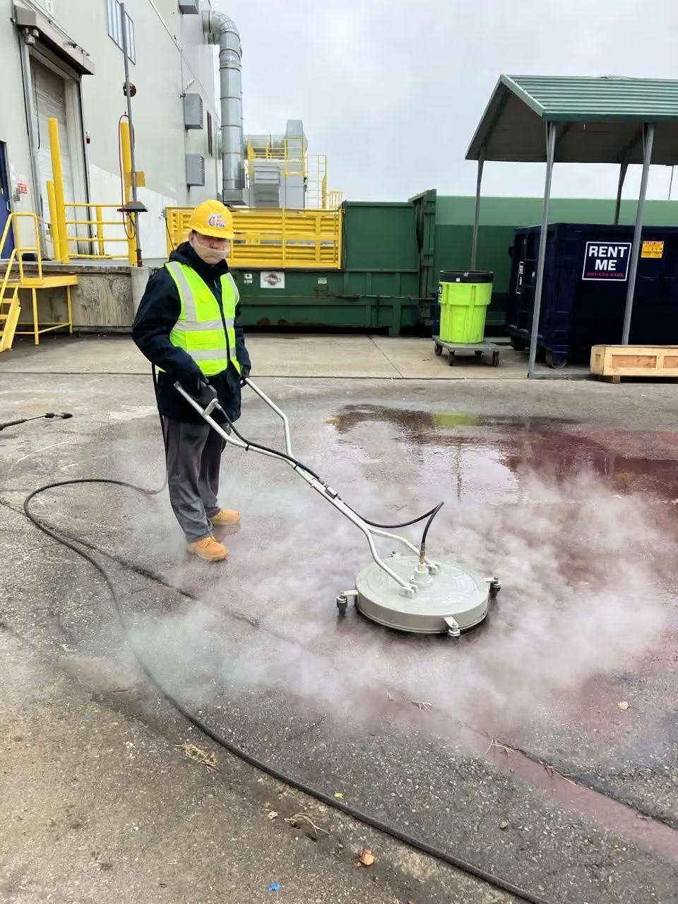 Worker pressure-washing pavement beside industrial tanks, wearing a yellow hard hat and high-vis vest