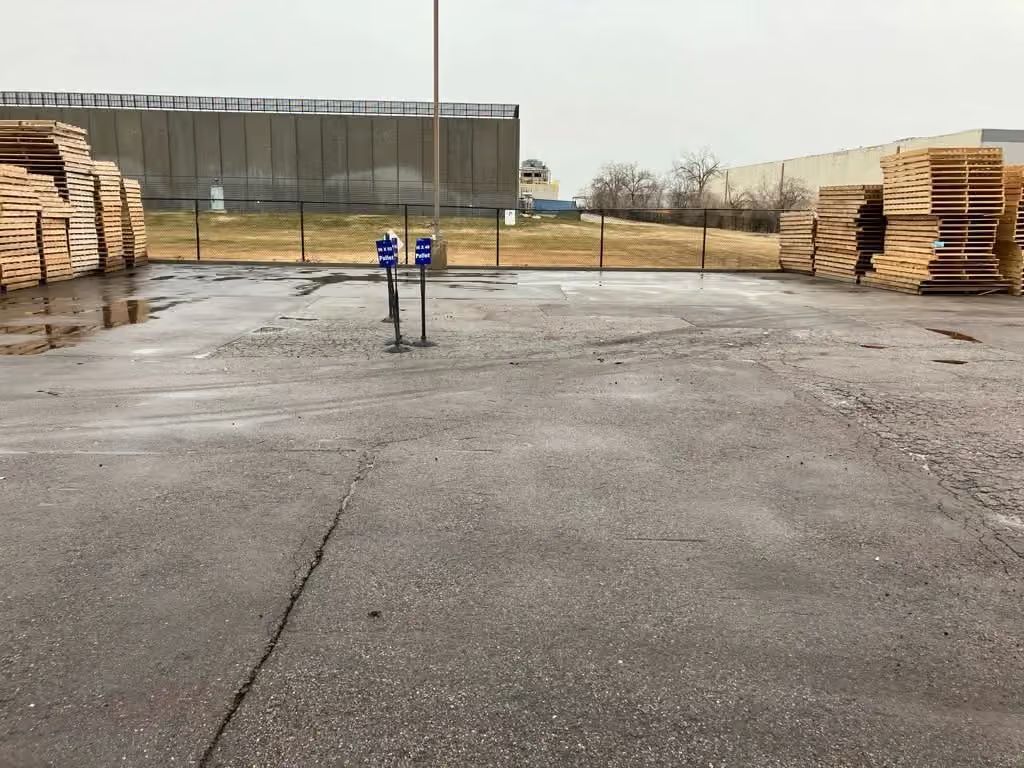 Wet industrial yard with stacked lumber and a gated fence in the background