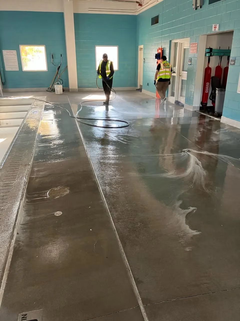 Wet gym floor in a turquoise-walled room with two workers wearing safety vests near the back.