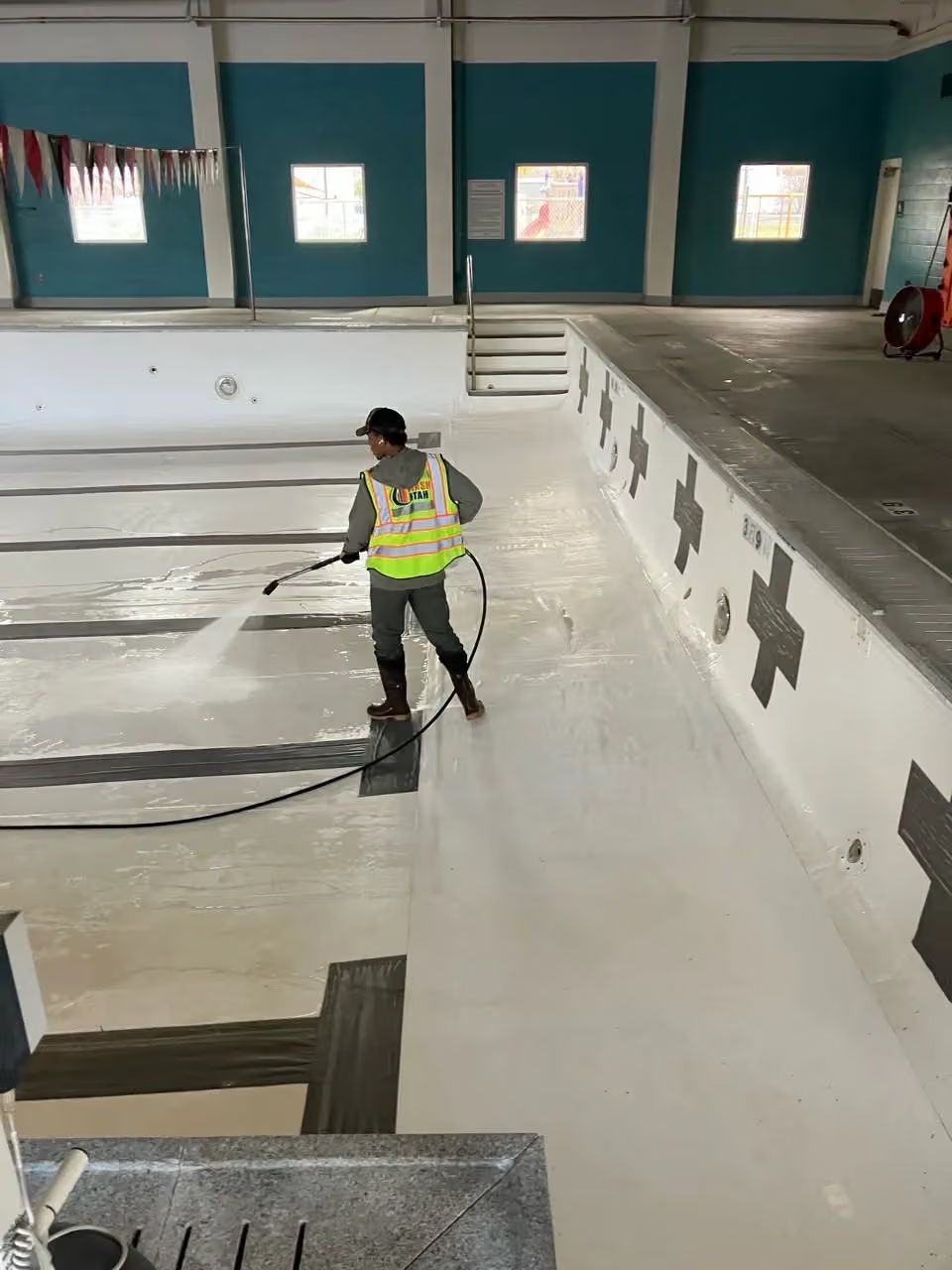 Worker in a reflective vest cleaning an empty indoor pool with a hose.