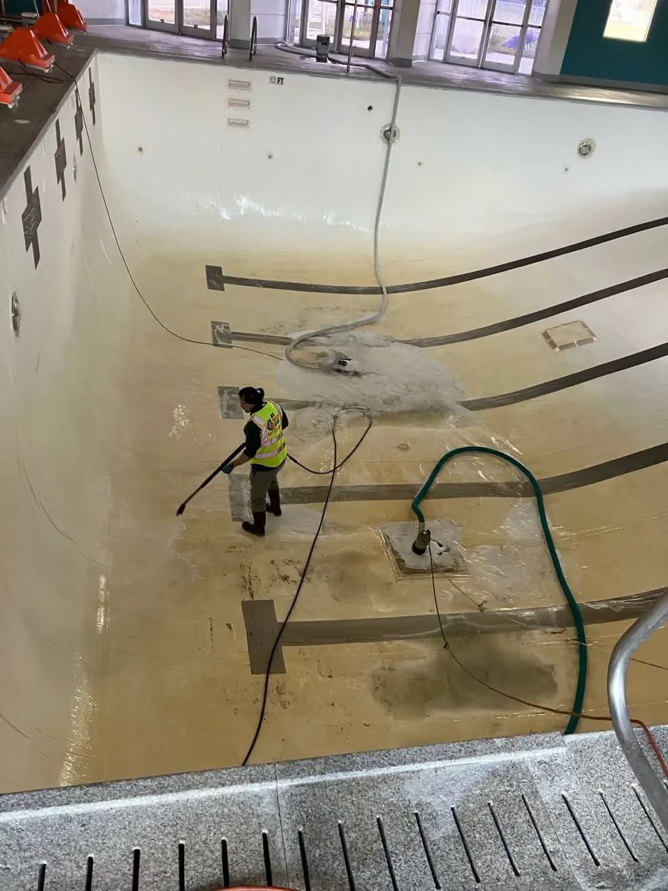 Person pressure washing an empty indoor swimming pool with hoses and cleaning equipment.