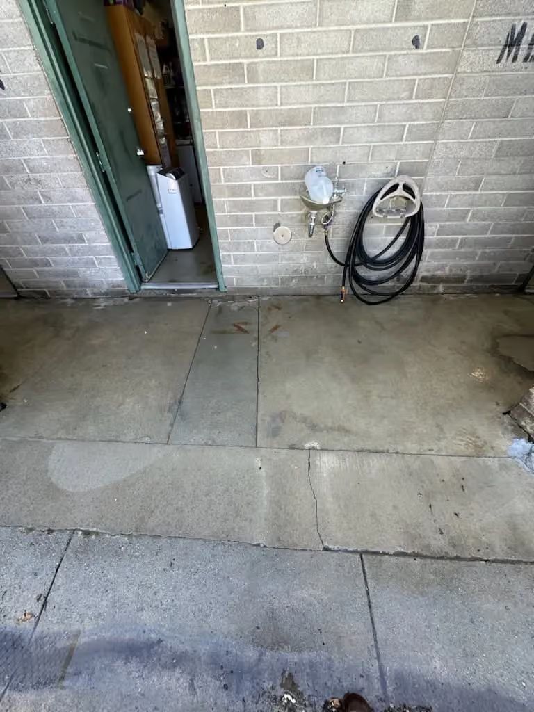 Concrete walkway beside brick wall with a doorway and coiled hose on an outdoor utility area.