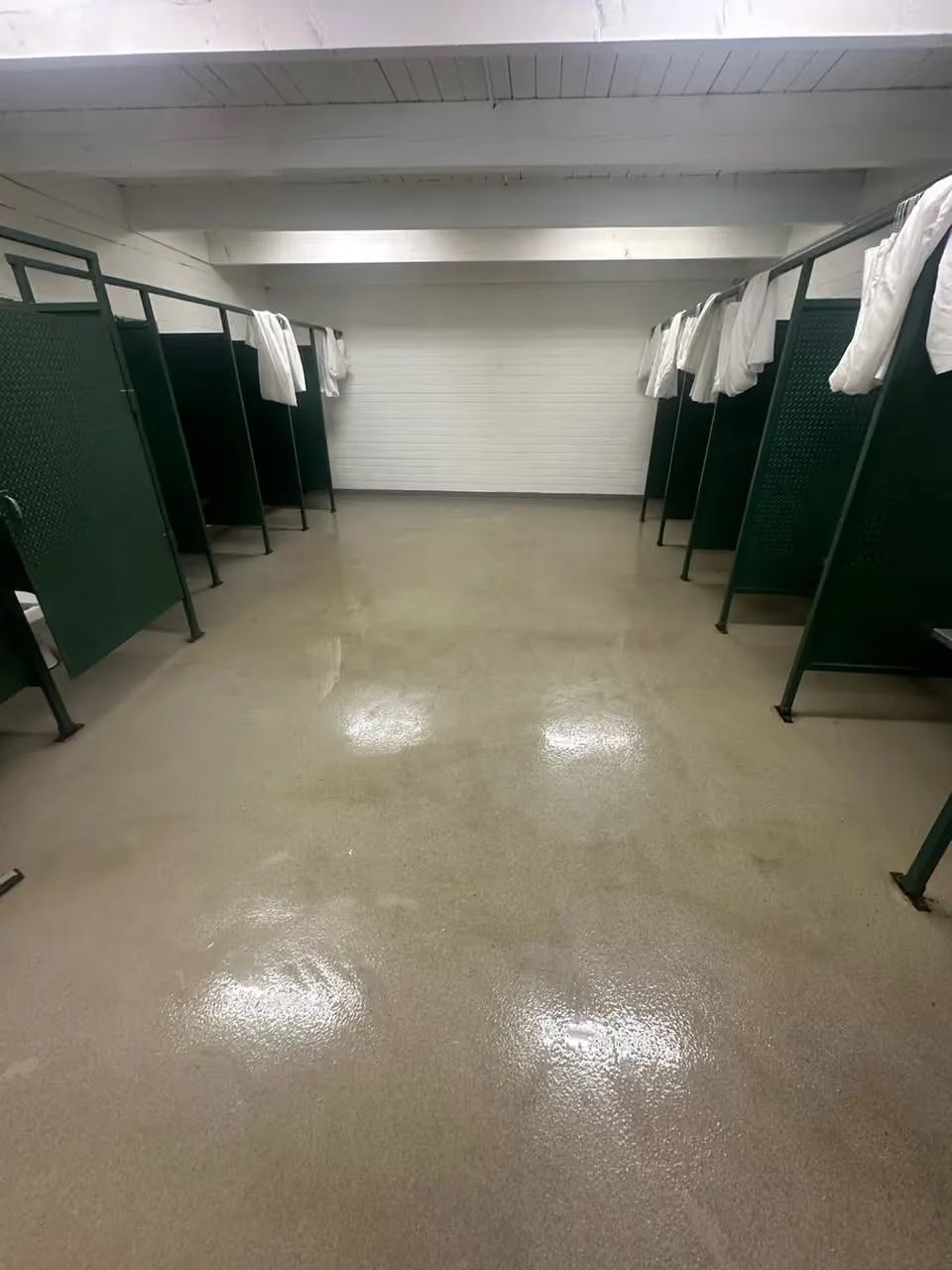 Empty locker room with green shower stalls and a glossy floor, viewed down the center aisle
